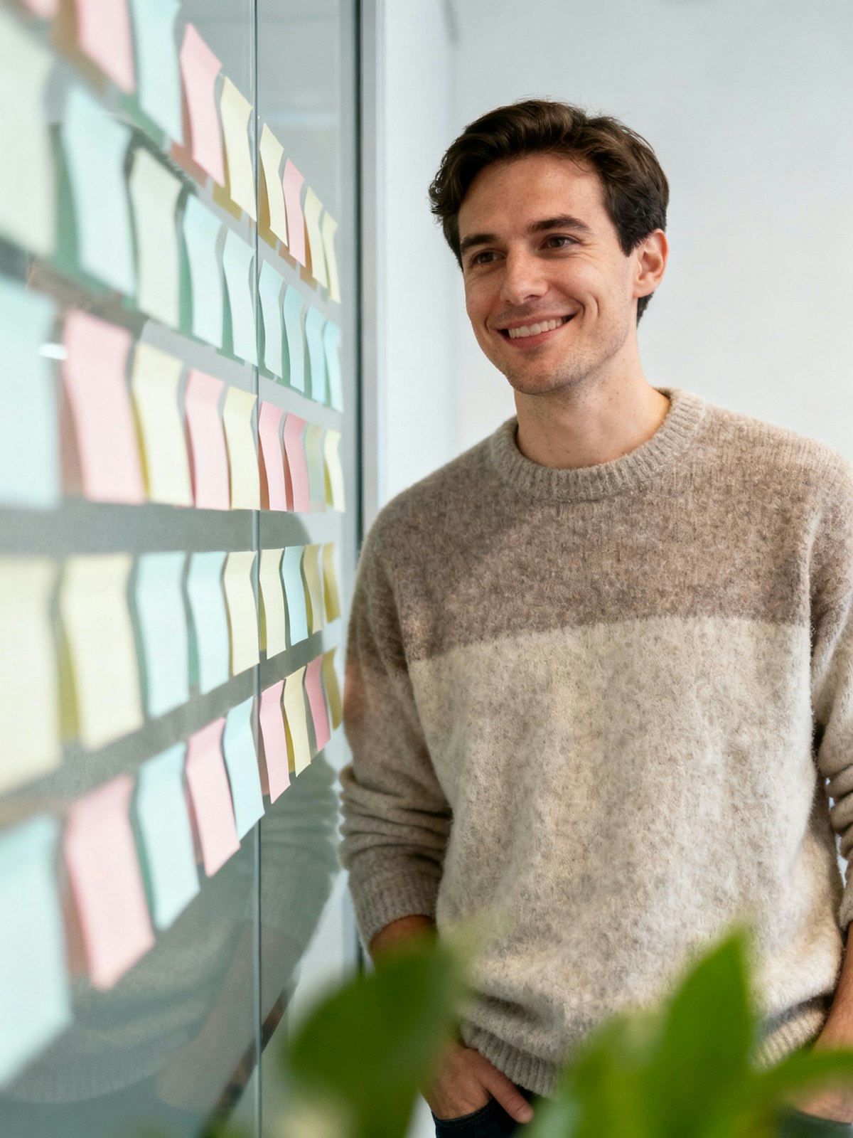 A smiling man in a grey sweater looking at rows of pastel sticky notes on a glass wall